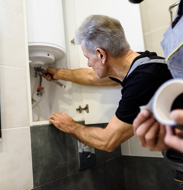 Affordable repairing. Aged plumber, repairman in uniform fixing electric boiler with wrench while his young colleague helping him, holding sticky duct tape indoors. Repair service concept Aged plumber, repairman in uniform fixing electric boiler with wrench while his young colleague helping him, holding sticky duct tape indoors. Repair service concept. Focus on person. Horizontal shot