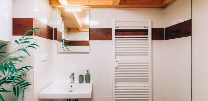 Bathroom wall with white washbasin and chrome faucet. On the wall there is mirror with bright glowing light and next to it is heating ladder. On the left is partly visible boiler and decorative plant.